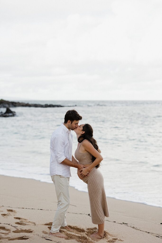 A pregnant woman kisses a man as they both hold the baby belly while standing on a beach in Hawaii