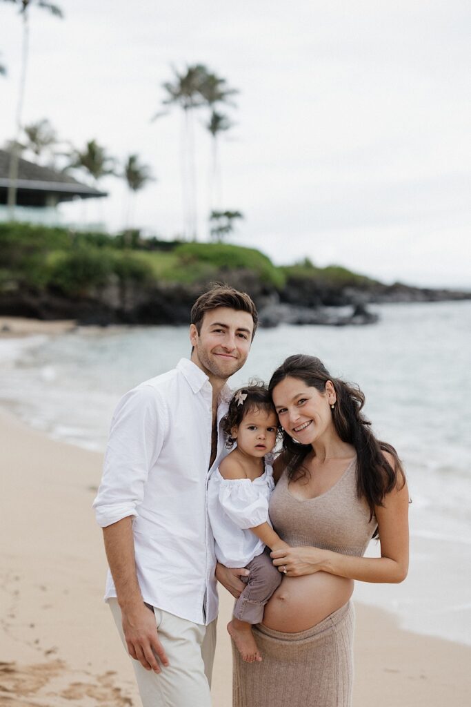 A father and pregnant mother smile at the camera while the mother holds their young daughter as they stand on a beach in Hawaii together
