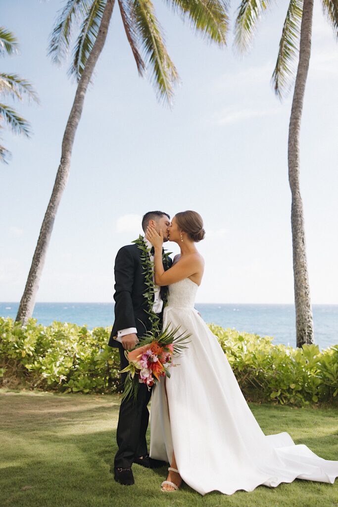 A bride and groom stand in a grass clearing and kiss one another with the ocean behind them 