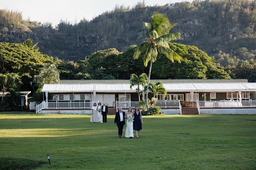 2 brides each escorted by their parents walk towards the camera for their wedding ceremony at Loulu Palm on Oahu
