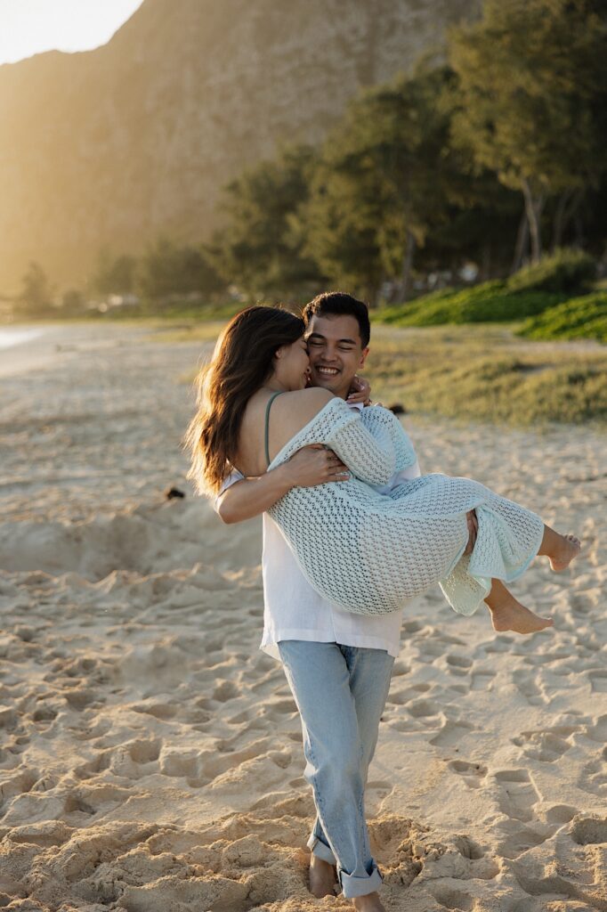A man smiles while carrying a woman in his arms along Waimanalo Beach on Oahu as the sun rises behind them
