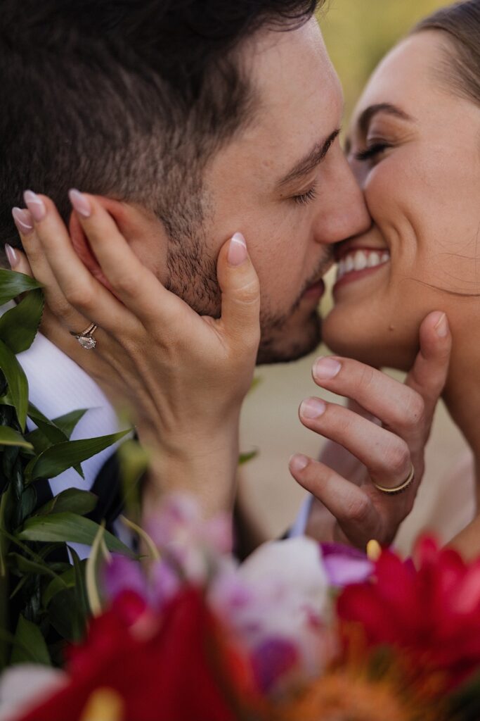 Close up photo of a bride and groom smiling as they're about to kiss one another, each is showing off their hand with their wedding ring on it