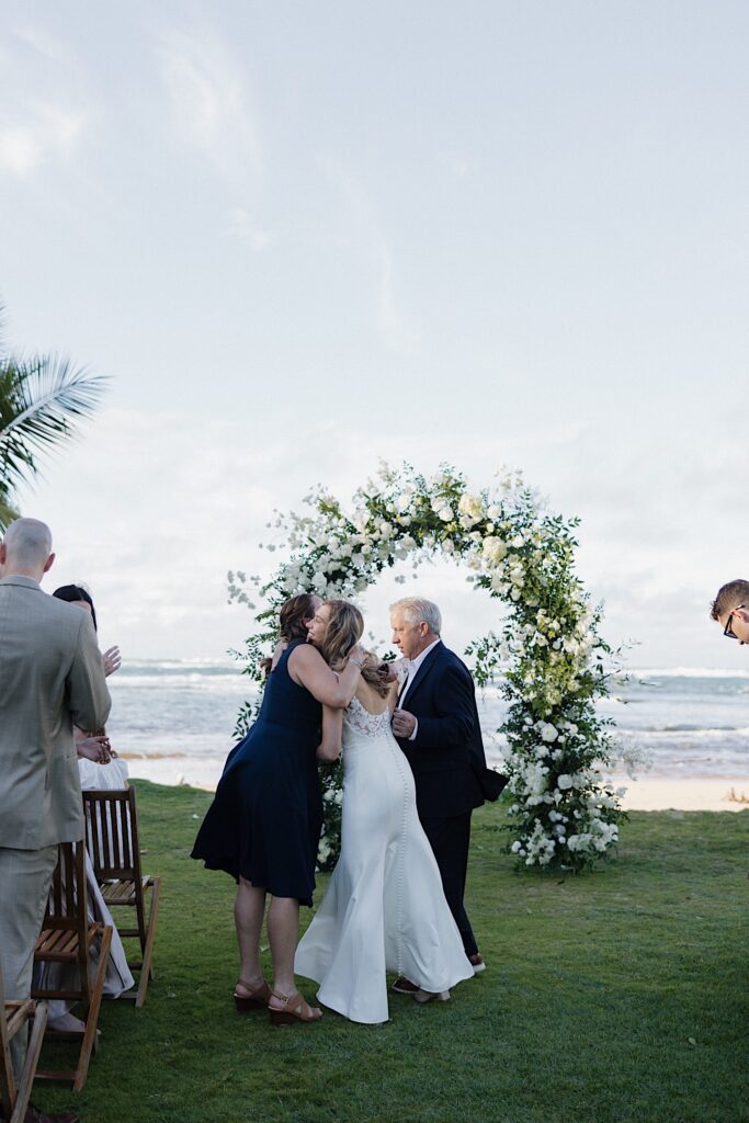 A bride is hugged by her parents in front of a floral altar that stands in front of the ocean at her wedding venue, Loulu Palm on Oahu