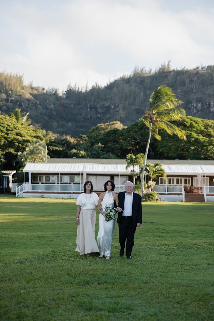 A bride is escorted by her parents towards her outdoor wedding ceremony at Loulu Palm on Oahu