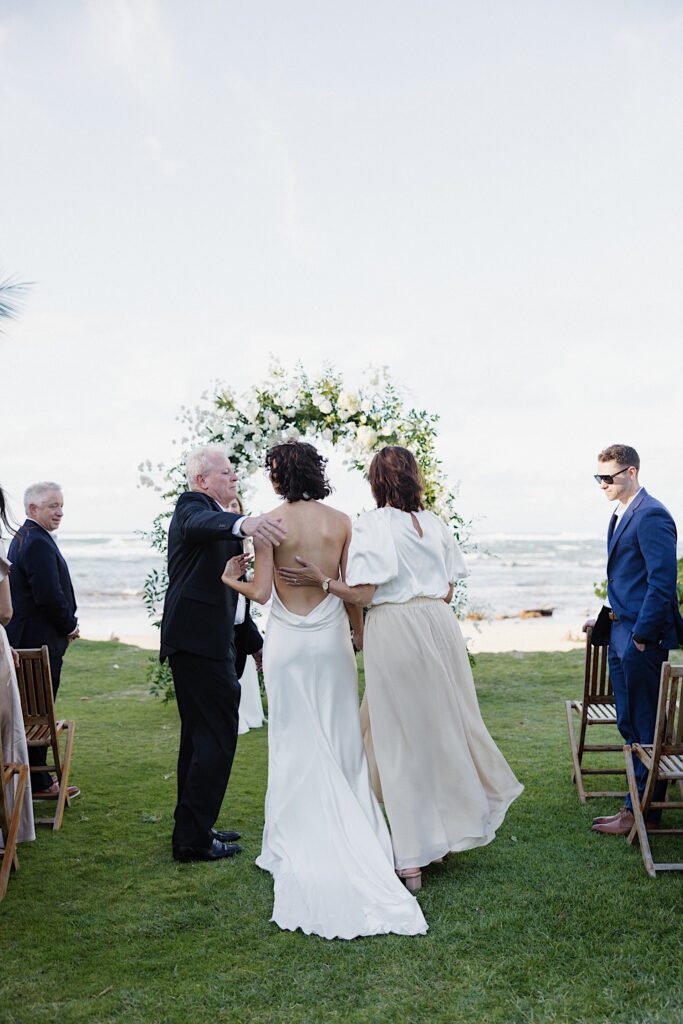 A bride is hugged by her parents while standing at her outdoor ceremony space in front of the ocean at Loulu Palm on Oahu