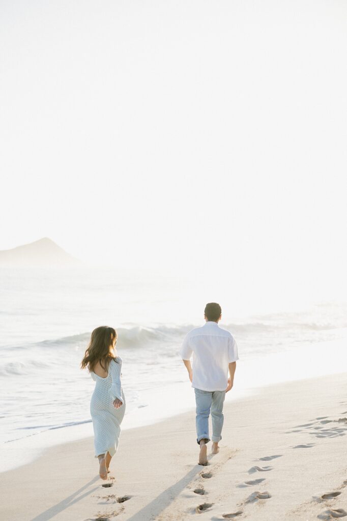 A couple walk along Waimanalo Beach on Oahu towards the water and the bright sun as it rises in the distance, photo taken by an Oahu Engagement photographer
