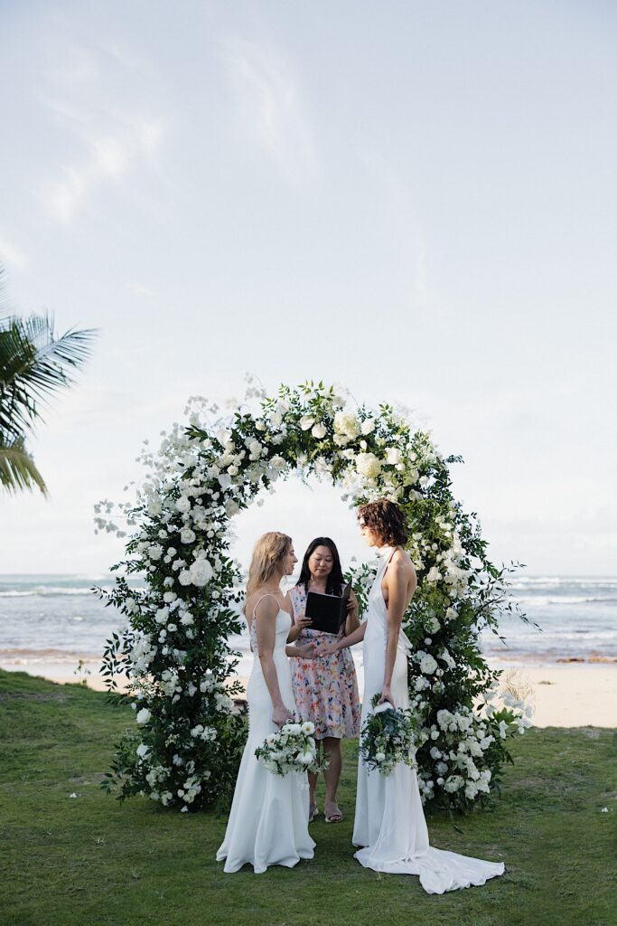 2 brides stand facing one another while their officiant reads during their outdoor wedding ceremony at Loulu Palm in front of the ocean