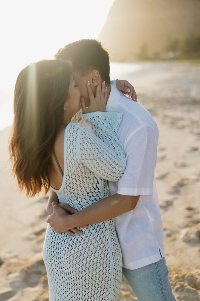 A couple hug and the woman kisses the man's cheek while the sun rises behind them over Waimanalo Beach