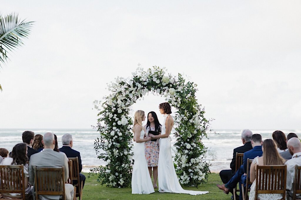 2 brides face on another and hold hands during their LGBTQ wedding ceremony at Loulu Palm on Oahu. Behind them is their floral altar, the ocean, and their officiant.