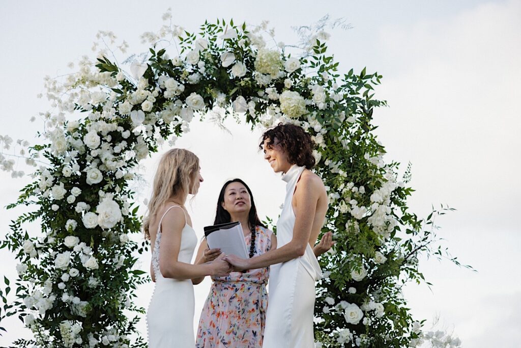 2 brides hold hands and smile towards one another while their officiant speaks standing under their floral altar during their LGBTQ wedding ceremony at Loulu Palm on Oahu