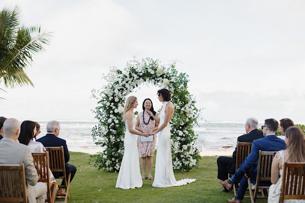 2 brides hold hands while one reads her vows during their LGBTQ wedding ceremony at Loulu Palm on Oahu, behind them is their officiant, floral altar, and the ocean.