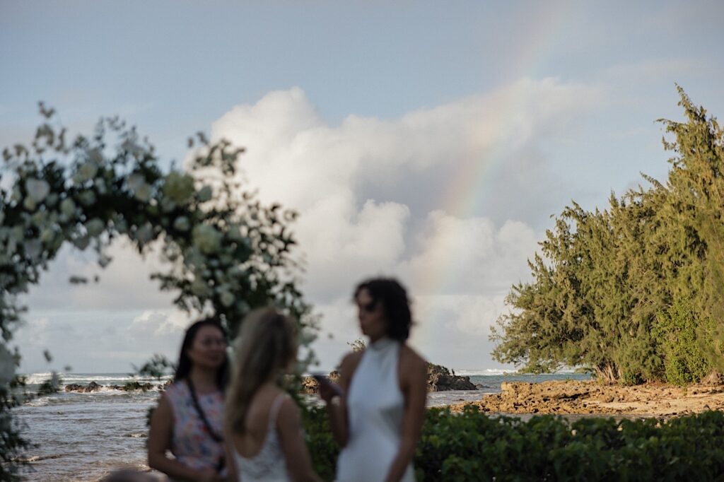 During an outdoor LGBTQ wedding ceremony at Loulu Palm on Oahu 2 brides stand in the foreground of the photo and face one another while a rainbow appears behind them