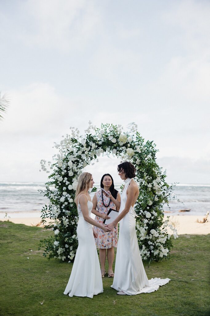 2 brides hold hands and face one another during their outdoor wedding ceremony at Loulu Palm, behind them is the ocean, their floral altar, and their officiant