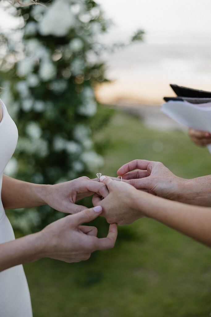 Close up photo of a bride putting a wedding ring on the other bride's hand during their outdoor wedding ceremony at Loulu Palm