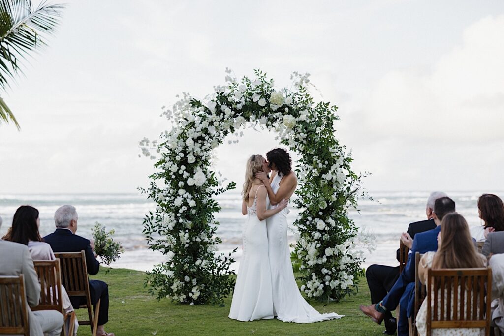 2 brides kiss one another while under a floral altar during their outdoor LGBTQ wedding ceremony at Loulu Palm on Oahu