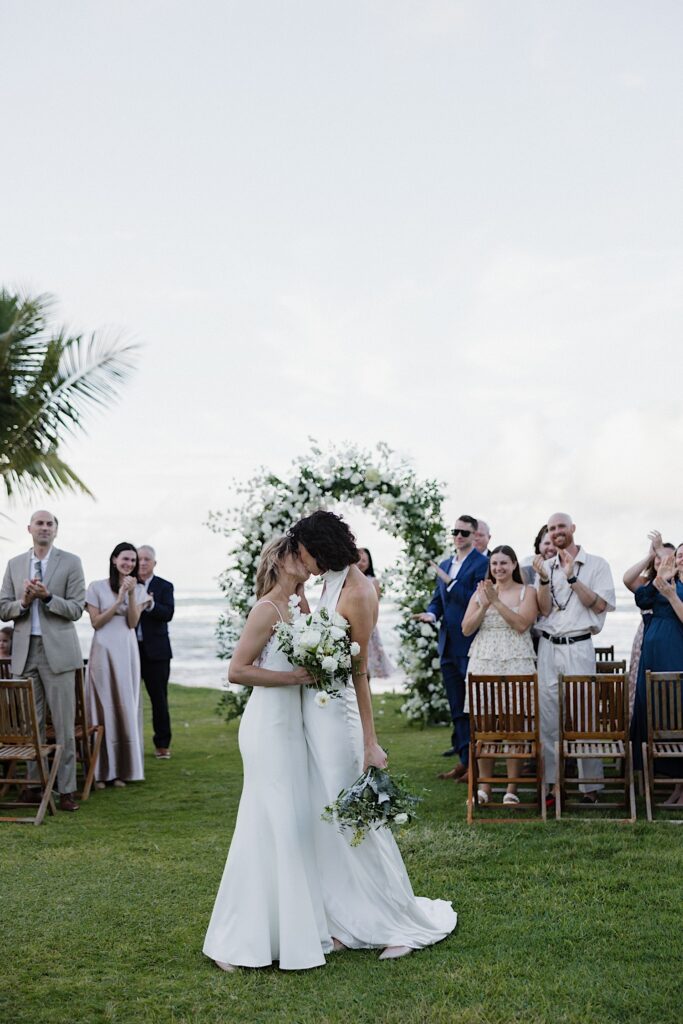 2 brides kiss one another as they exit their outdoor wedding ceremony at Loulu Palm while guests clap behind them
