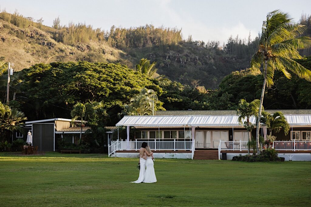 2 brides embrace while facing away from the camera in the distance as they stand in the lawn of their wedding venue, Loulu Palm on Oahu, after their LGBTQ wedding ceremony.