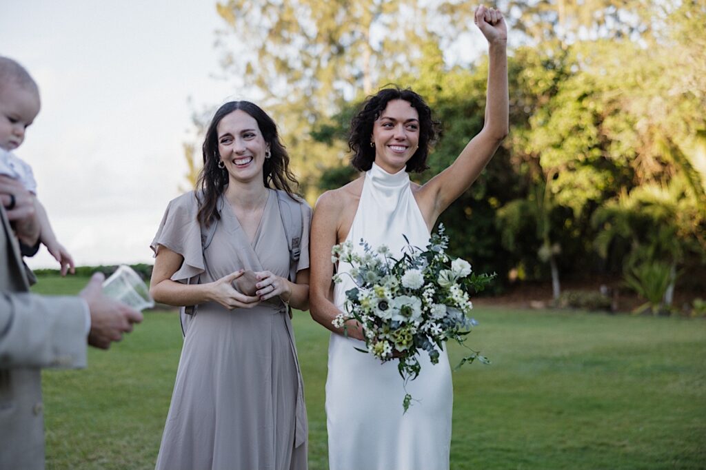 A bride smiles and lifts her arm in the air while standing beside a guest of her LGBTQ wedding day at Loulu Palm on Oahu