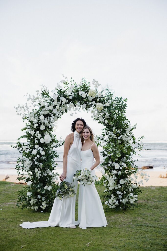 2 brides smile at the camera while standing side by side in front of the ocean and the floral altar of their outdoor ceremony space at Loulu Palm