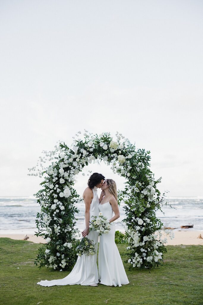 2 bride kiss one another while in front of their floral altar and the ocean at the ceremony space of Loulu Palm