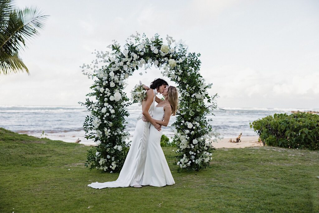 2 brides kiss one another while in front of the floral altar and the ocean at their LGBTQ wedding venue, Loulu Palm on Oahu
