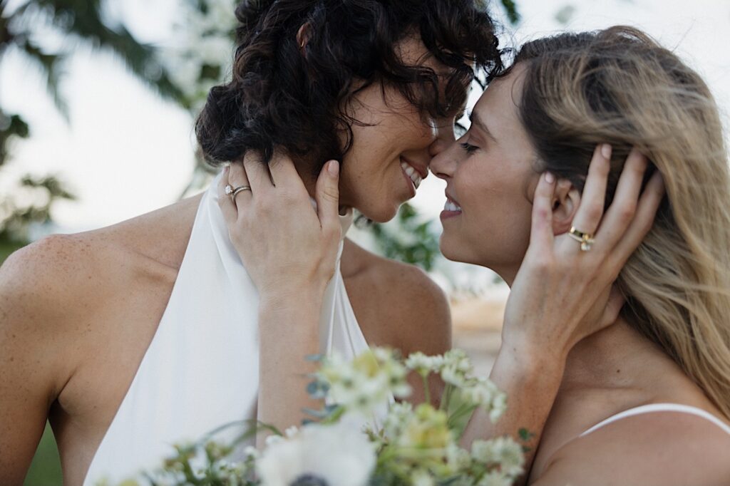 2 brides hold one another and smile as they are about to kiss after their outdoor LGBTQ wedding at Loulu Palm on Oahu