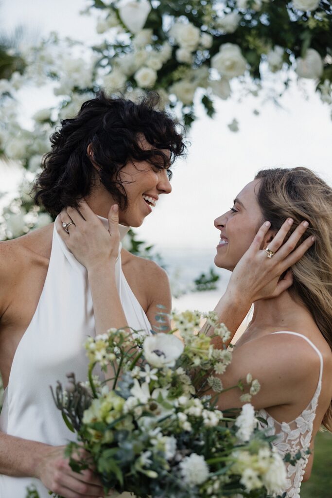 2 brides smile at one another while holding each other's faces underneath a floral arch at their venue, Loulu Palm