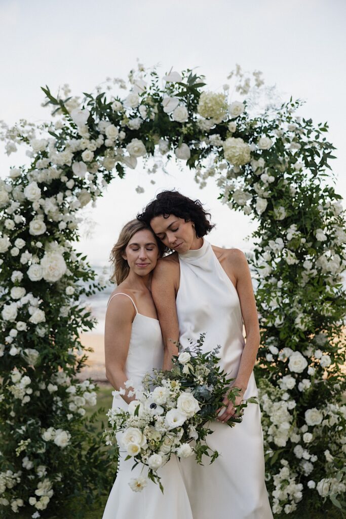 2 brides stand side by side and close their eyes while under a floral arch at their wedding venue Loulu Palm