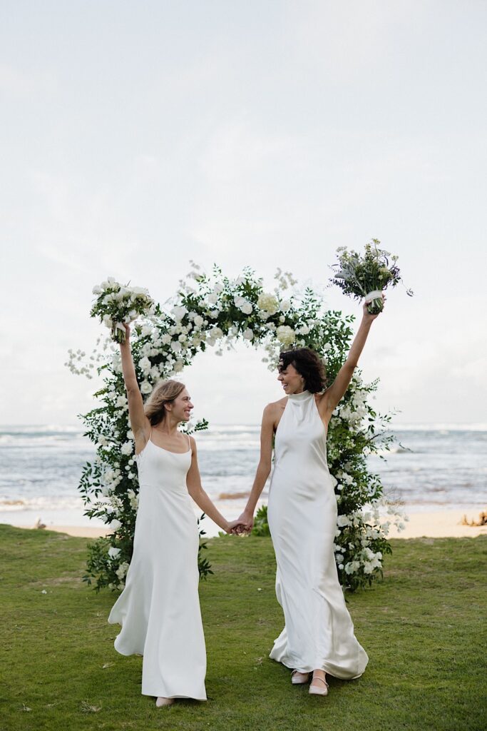 2 brides hold hands, smile at one another, and raise their bouquets in the air while walking towards the camera at the outdoor ceremony space at Loulu Palm