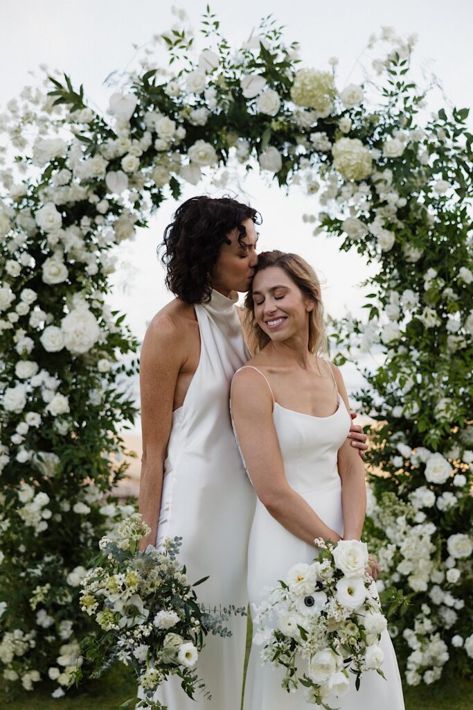 A bride smiles while the bride behind her kisses her head as they stand underneath a floral altar