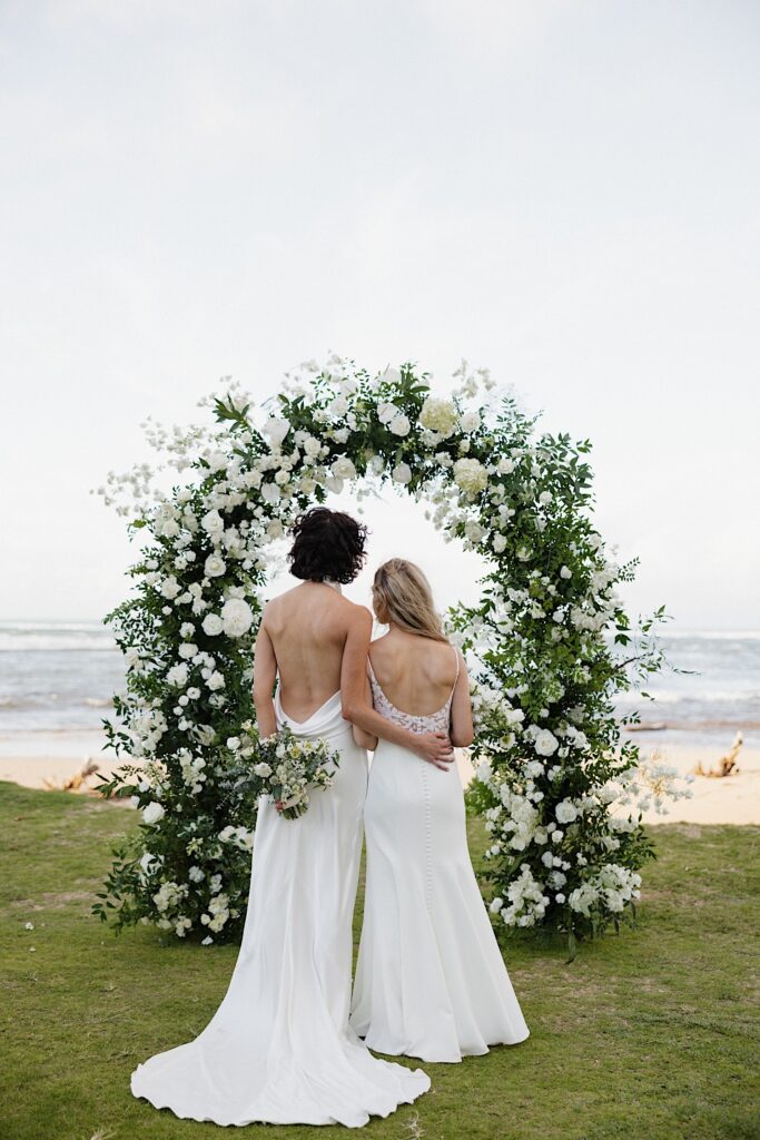 2 brides face away from the camera towards their floral altar and the ocean in the background at Loulu Palm on Oahu