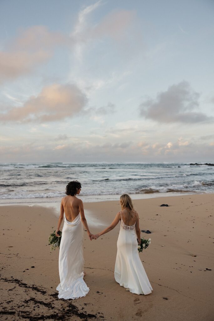 2 brides hold hands and walk away from the camera on the beach towards the ocean at sunset