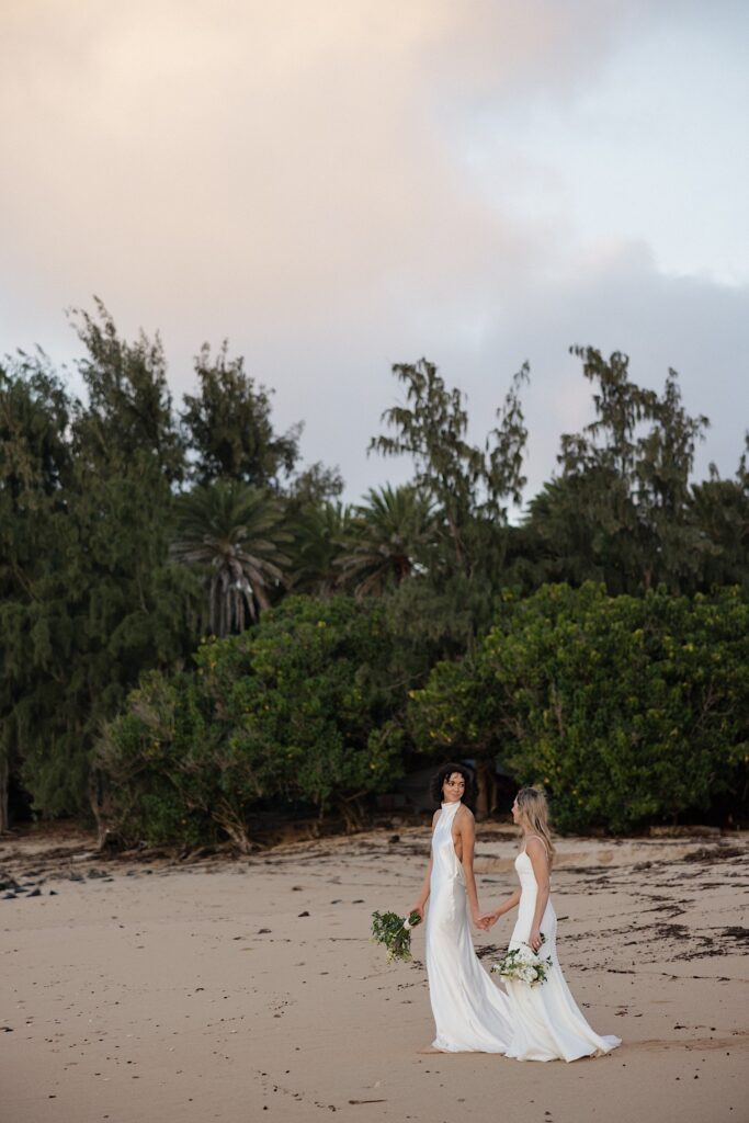 2 brides walk along the beach while holding hands and looking at one another during sunset near their venue Loulu Palm on Oahu