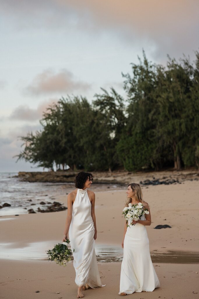 2 brides walk along the beach and smile towards one another as the sun sets near their wedding venue Loulu Palm