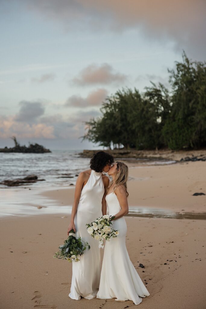 2 brides kiss one another while on a beach of Oahu near Loulu Palm as the sun sets