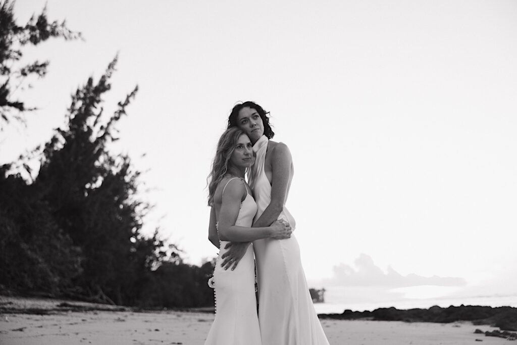 Black and white photo of 2 brides holding one another while standing on a beach of Oahu near the venue for their LGBTQ wedding, Loulu Palm