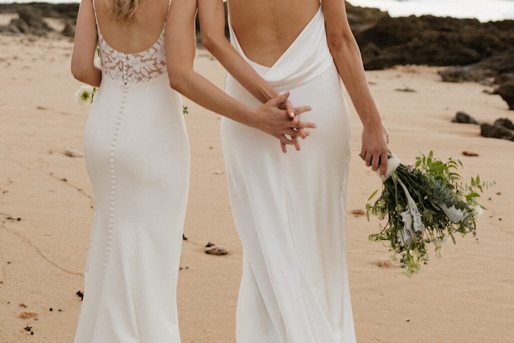 Close up photo of two brides holding hands behind their backs as they walk along the beach near the venue for their LGBTQ wedding, Loulu Palm on Oahu