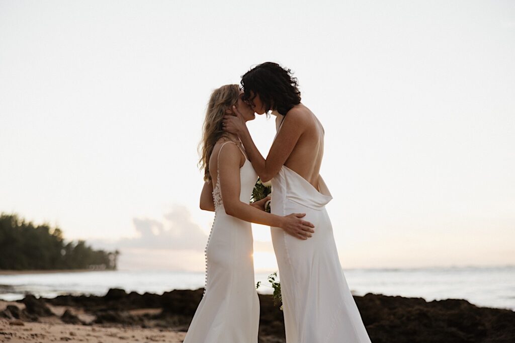 2 brides stand on a beach at sunset as they kiss one another after their LGBTQ wedding ceremony at Loulu Palm on Oahu