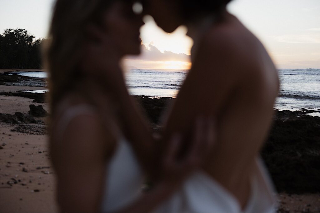 2 brides embrace one another in the foreground as the sun sets behind them while they're on a beach near the venue for their LGBTQ wedding, Loulu Palm on Oahu