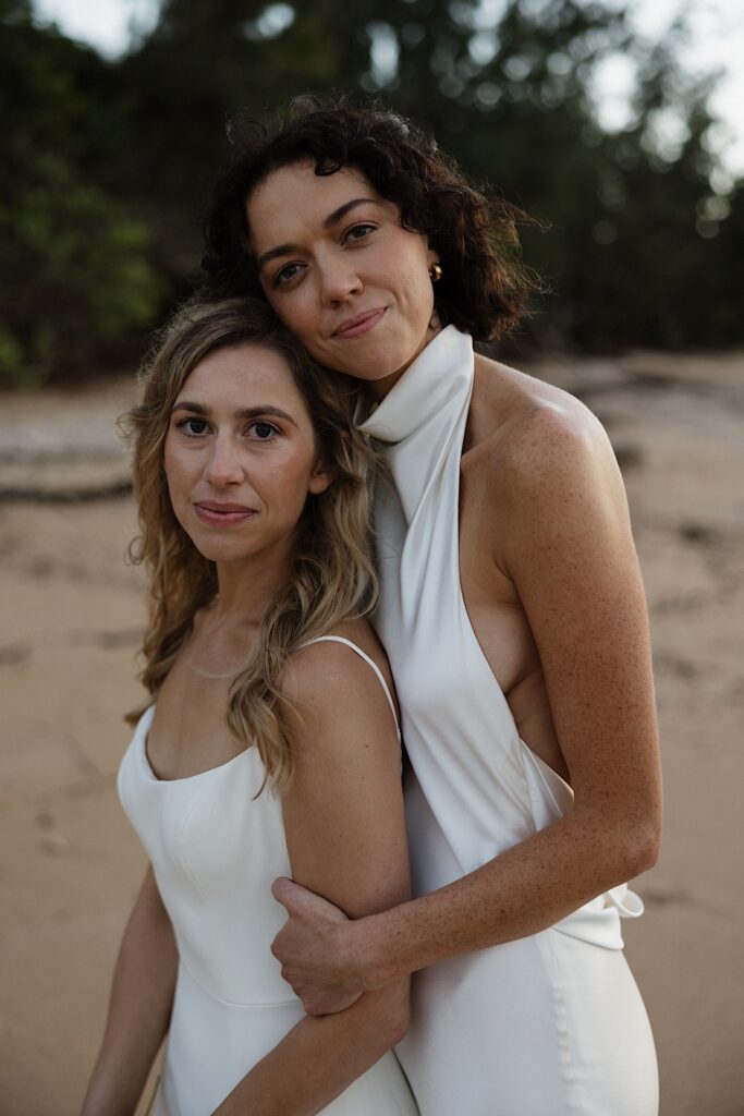 2 brides look at the camera, one hugging the other from behind, while standing on a beach of Oahu