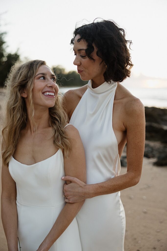 2 brides smile at one another, one standing behind the other, while standing on a beach of Oahu
