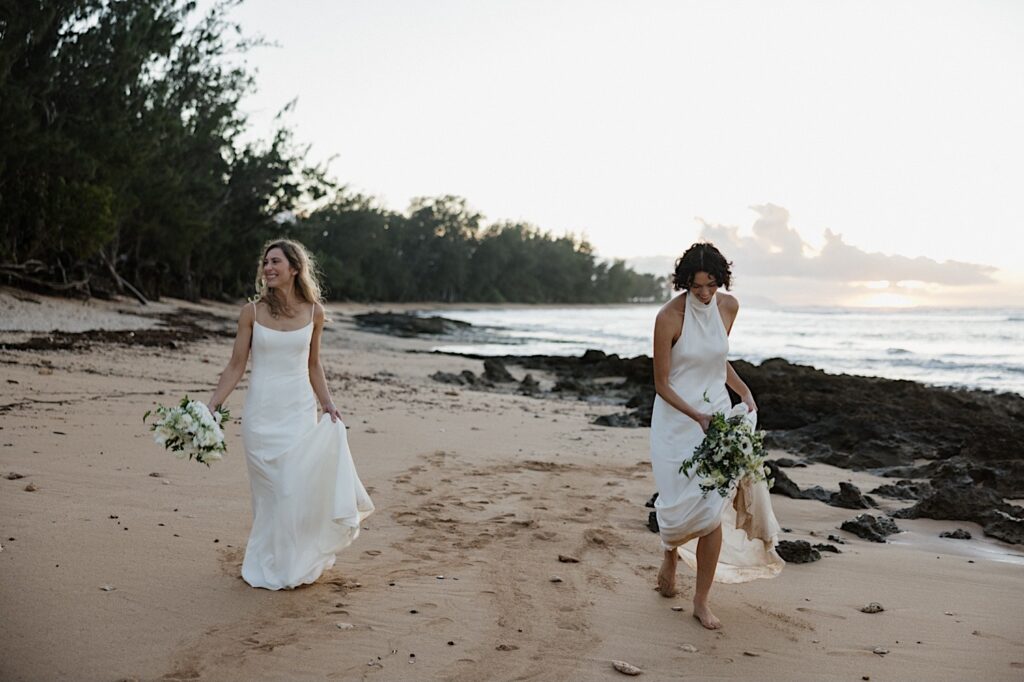2 brides walk along a beach of Oahu near the venue of their LGBTQ wedding, Loulu Palm, while the sun sets on the ocean behind them