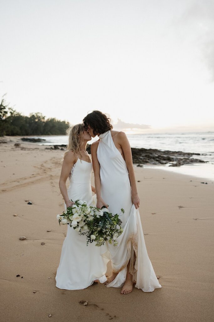 2 brides kiss one another while standing on a beach of Oahu as the sun sets on the ocean behind them
