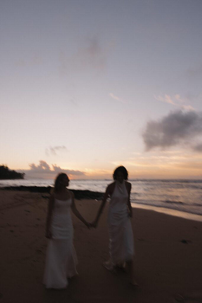 2 brides hold hands and walk towards the camera while on a beach of Oahu at sunset