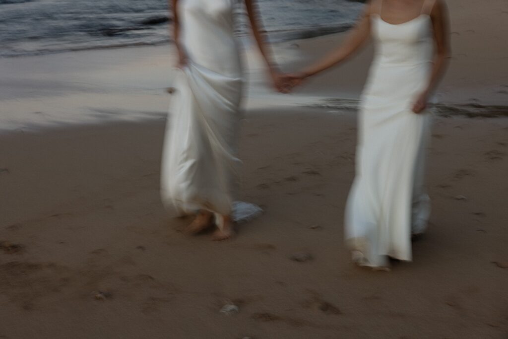 2 brides hold hands and walk along a beach of Oahu near the venue of their LGBTQ wedding, Loulu Palm, at sunset