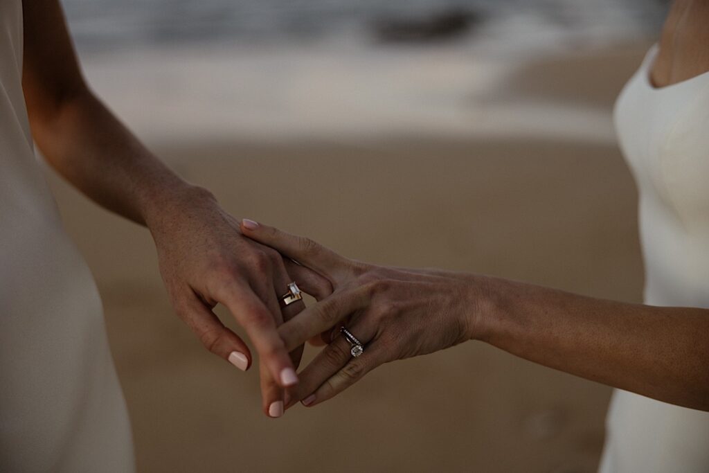 Close up photo of two bride's hands with their wedding rings holding one another as they stand on the beach near their LGBTQ wedding venue, Loulu Palm on Oahu