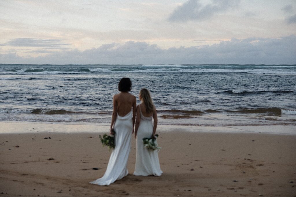 2 brides stand site by side on a beach near their LGBTQ wedding venue, Loulu Palm on Oahu, and look out at the ocean at sunset