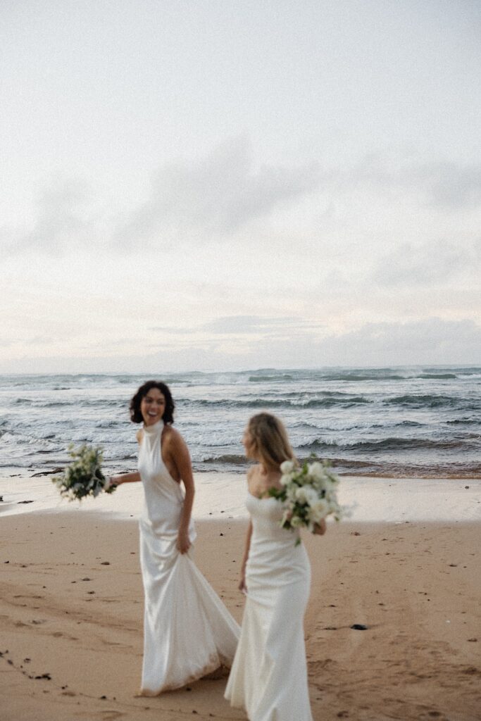 2 brides smile at one another while walking along a beach on Oahu at sunset with the ocean behind them