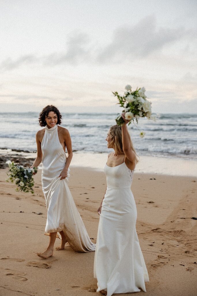 2 brides smile at one another while walking along a beach on Oahu at sunset with the ocean behind them