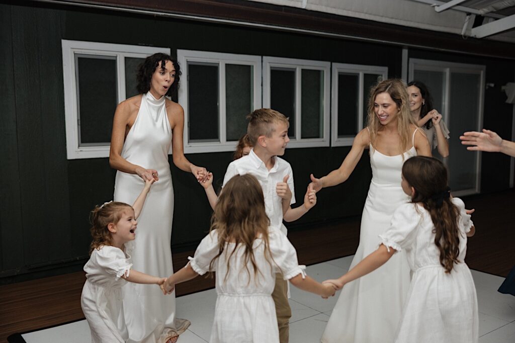 2 brides dance in a circle with children at their outdoor LGBTQ wedding reception at Loulu Palm on Oahu